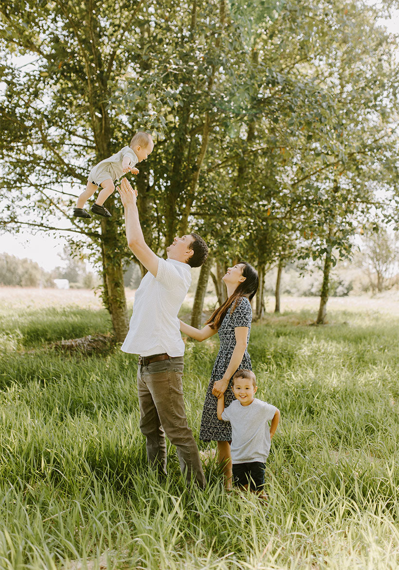 The family behind Wholesome Story outdoors under trees: a father lifts a baby into the air, and a mother and a young boy look on